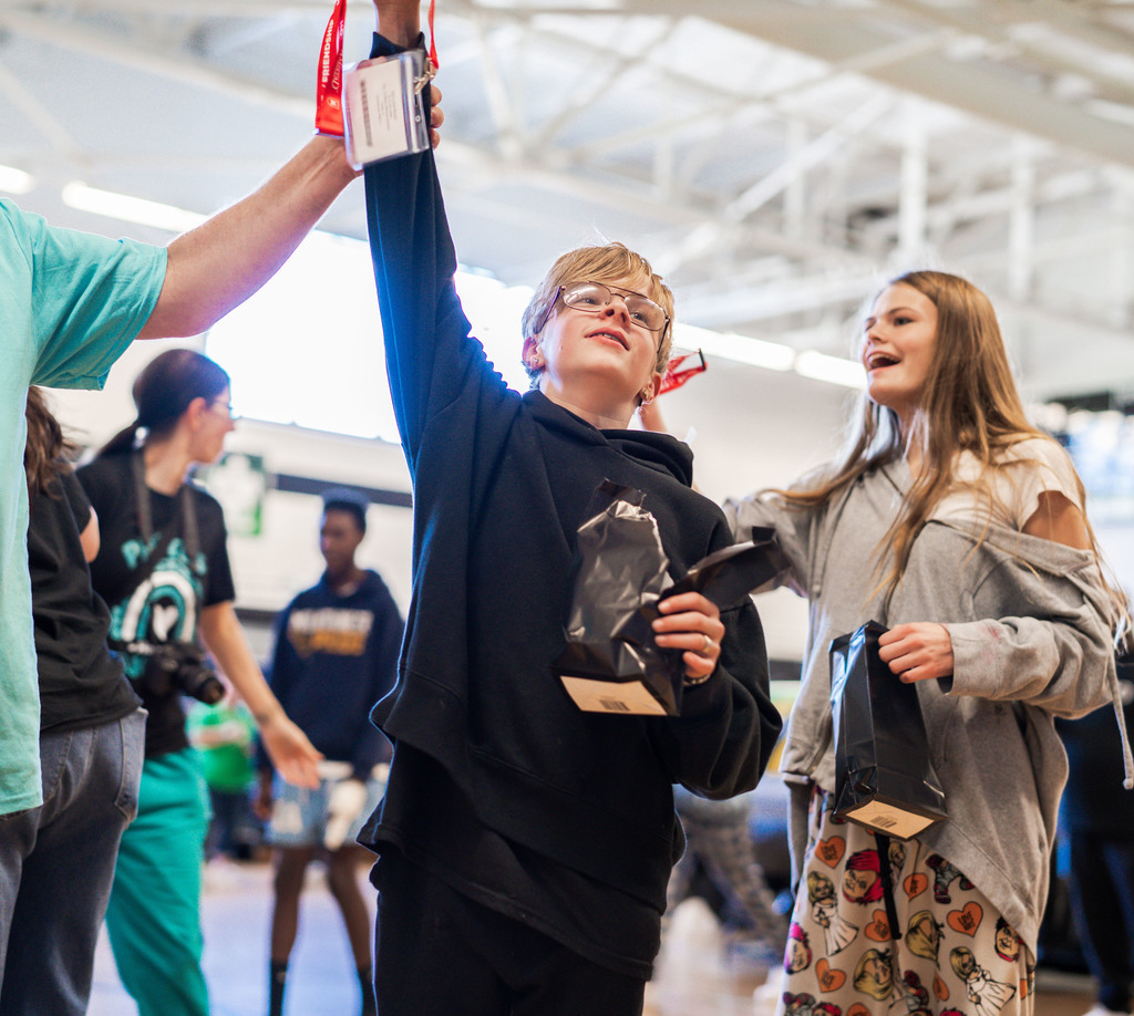 student holding his hand up to show off a lanyard he received. Behind him, another student is showing off her lanyard to a friend that is not in the picture.
