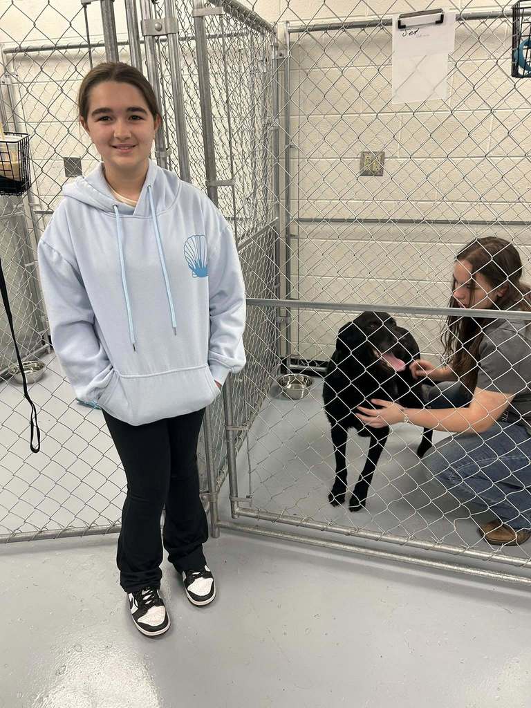 A nitro student is standing in front of a dog kennel at the ben franklin grooming class. In the kennel is a black dog, breed unknown, and a ben franklin student it petting the dog. 
