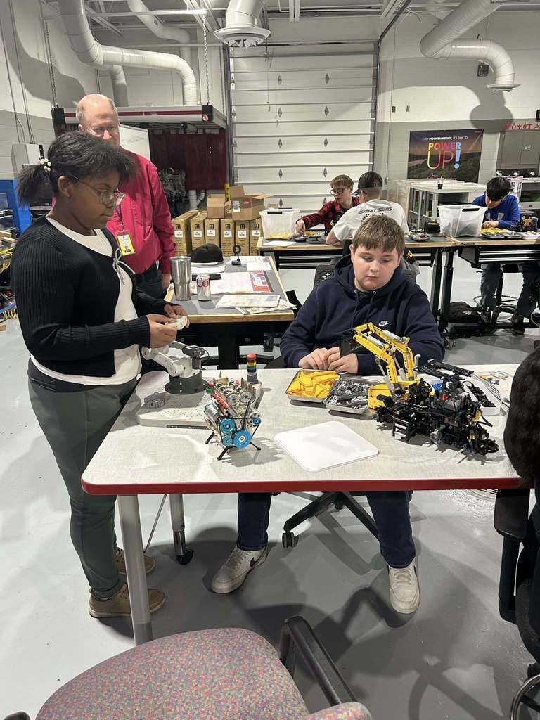 one nitro student is sitting at a workshop table at ben franklin while another is standing at the side of the table. On the table are  different types of mini engines and equipment. 