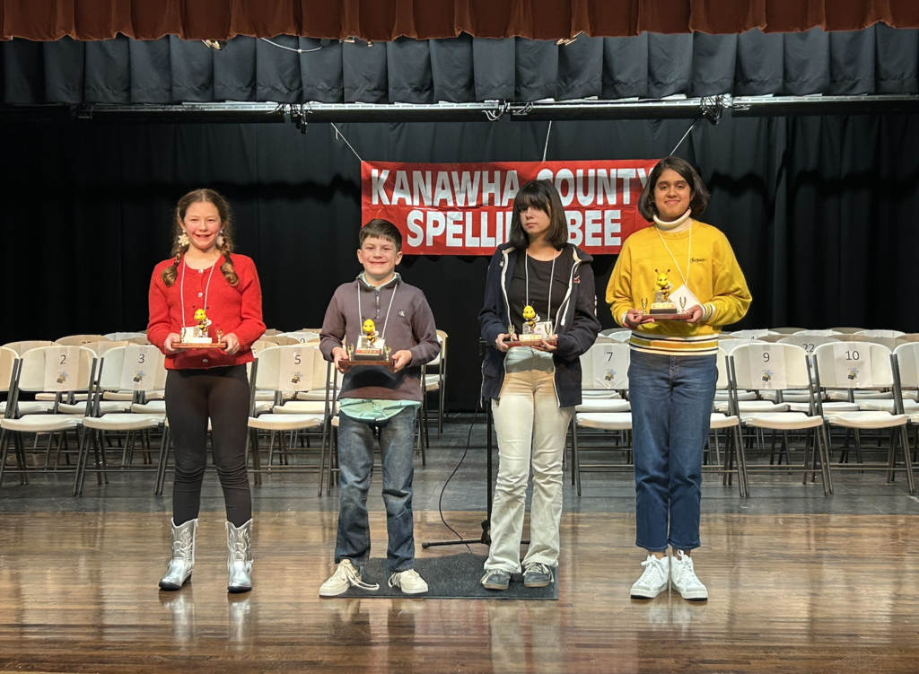 Four students on stage at the KCS spelling bee. All four students are holding trophies for placing first or runner up in the KCS spelling bee. Two from the Middle School contest and Two from the Elementary contest.