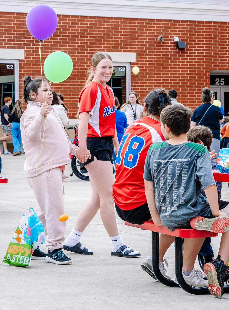 A Herbert Hoover student, wearing her red softball jersey, is holding hands with a younger student as they walk around the courtyard area during the event. The younger student is holding a purple balloon and a green balloon. Students sitting down at a near by table and looking at the two as they walk by.