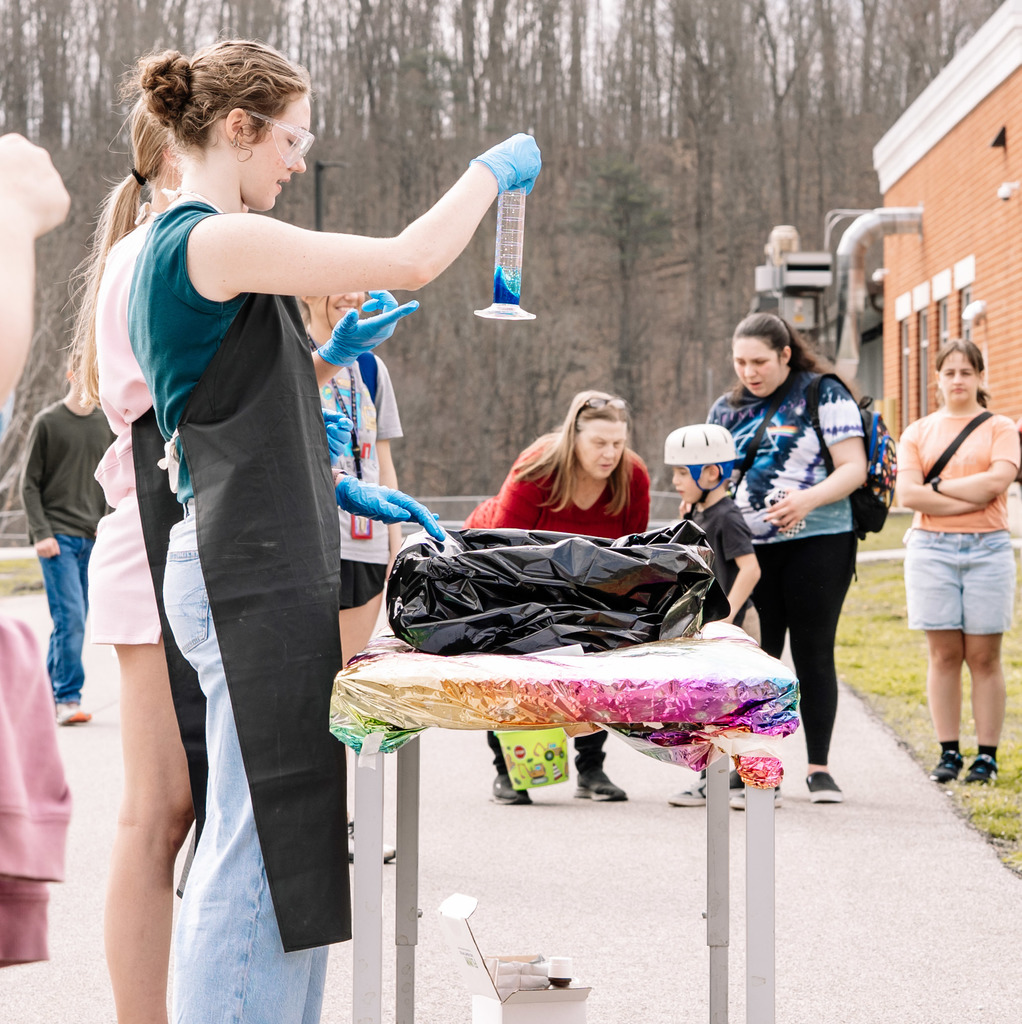 A Herbert Hoover student is holding up a graduated cylinder that has blue liquid in the bottom of it. The student is wearing blue latex gloves, a black lab apron, and has on safety goggles.