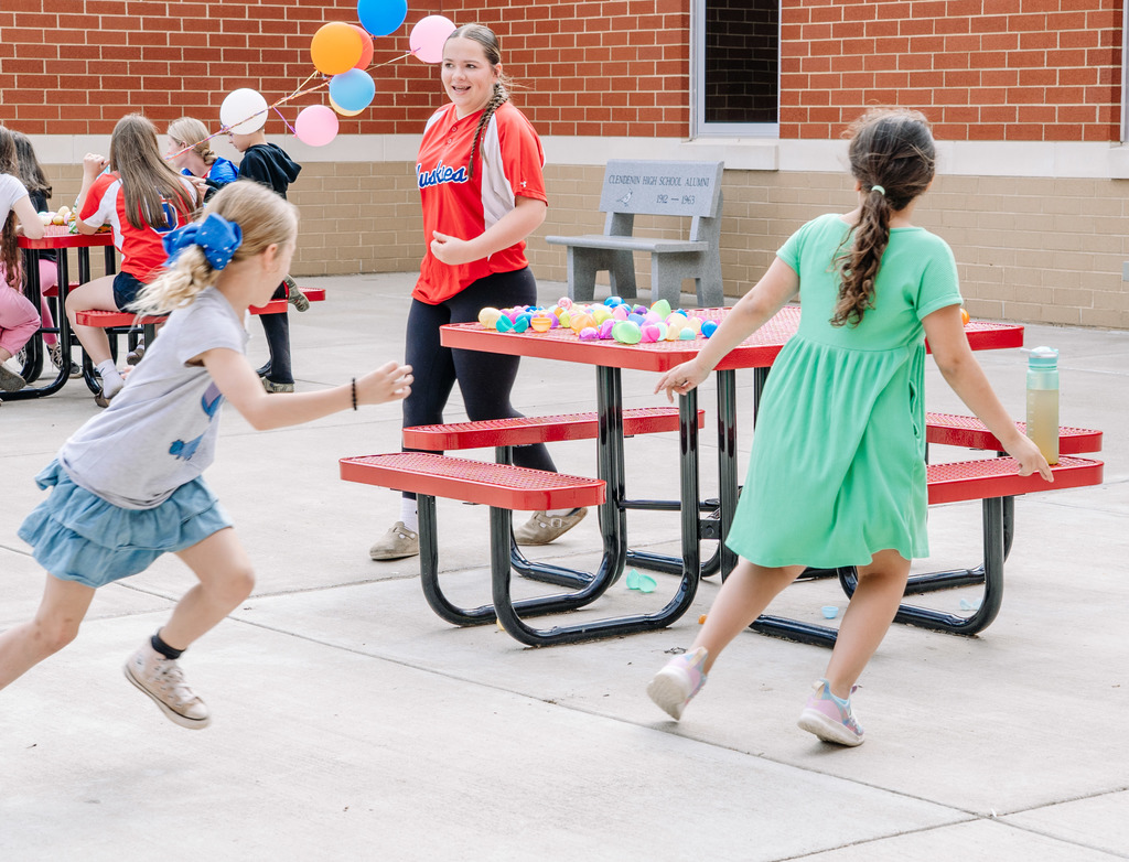 Two young students and a Herbert Hoover student are running around one of the red patio tables playing a game of tag. The Herbert Hoover student is chasing the two younger students.