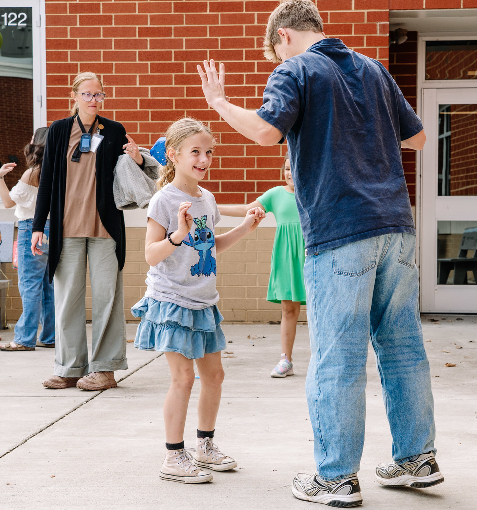 A young student, wearing a Lilo and Stich shirt, is playing tag with an older student. The younger student is starting to reach out to tag the older student, and the older student has both of his hands in the air to signal that he has been caught.
