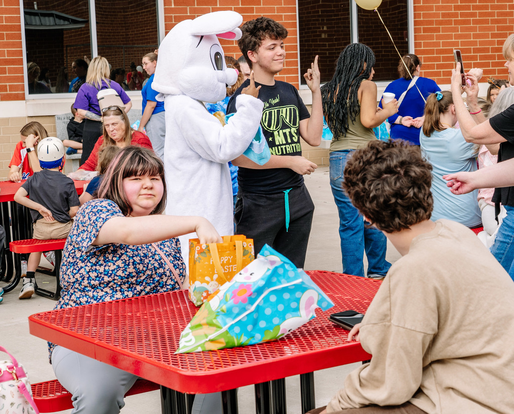 The Easter Bunny is posing for a picture with a student. They are making the rock and roll hand gesture with their index and pinkie fingers. In front of them, is a red patio table with two students sitting down. One student is looking at the easter bunny and the other is looking at the camera while reaching in her bag used for collecting the eggs.
