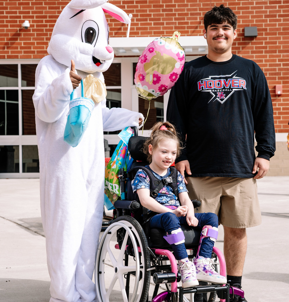 The easter bunny and a herbert hoover student are posing for a picture with a student in a wheelchair. The wheelchair has a pink and gold balloon with flowers on it tied to the back.