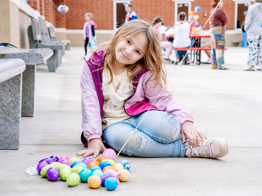 A young student is sitting down on the ground smiling at the camera. The easter eggs the student collected are sitting on the ground in front of her.