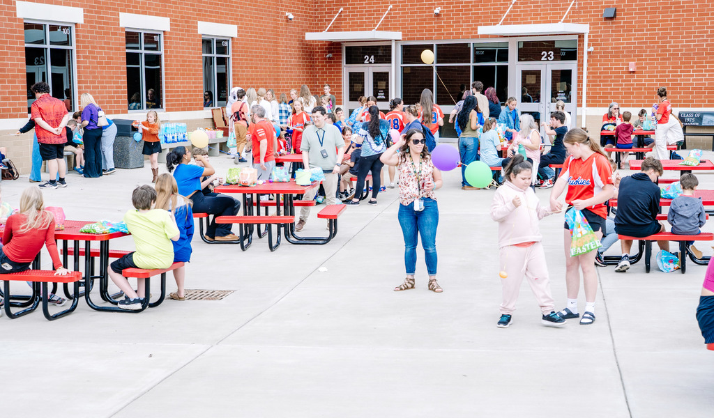 Students and teachers are scattered about in the HHHS courtyard during the Egg Hunt event. Some students are sitting at the patio tables and others are walking around.