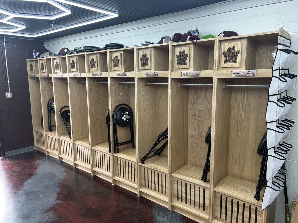 A row of lockers in the GWHS baseball locker room. There are stools for players to sit on, folded up, leaning against the inside wall of each locker. The stools are black and have the GW letters on them. On top of the lockers are cleats, hats, baseball gloves, and batting helmets.