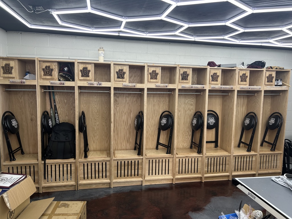 A row of lockers in the GWHS baseball locker room. There are stools for players to sit on, folded up, leaning against the inside wall of each locker. The stools are black and have the GW letters on them.