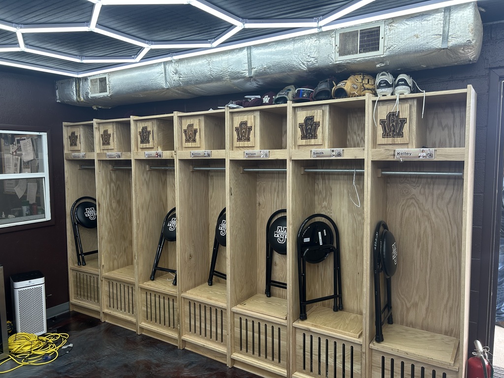A row of lockers in the GWHS baseball locker room. There are stools for players to sit on, folded up, leaning against the inside wall of each locker. The stools are black and have the GW letters on them. On top of the lockers are cleats, hats, baseball gloves, and batting helmets.