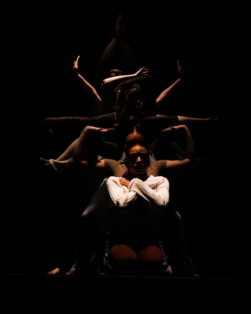 Dancers, some sitting and some standing, are in a straight line. The first dancer in the line has her arms crossed and her hands on her shoulders. Behind her, the rest of the dancers are making different lines and shapes with their arms. The picture is dark and moody. 
