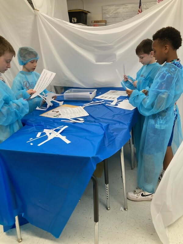 Four students, all wearing surgical gowns, are standing at their desks that have been pushed together and covered with a blue plastic table cloth to resemble an operating table. A white sheet is hanging  up around the students to create a makeshift operating room. The students are all cutting paper with scissors. 