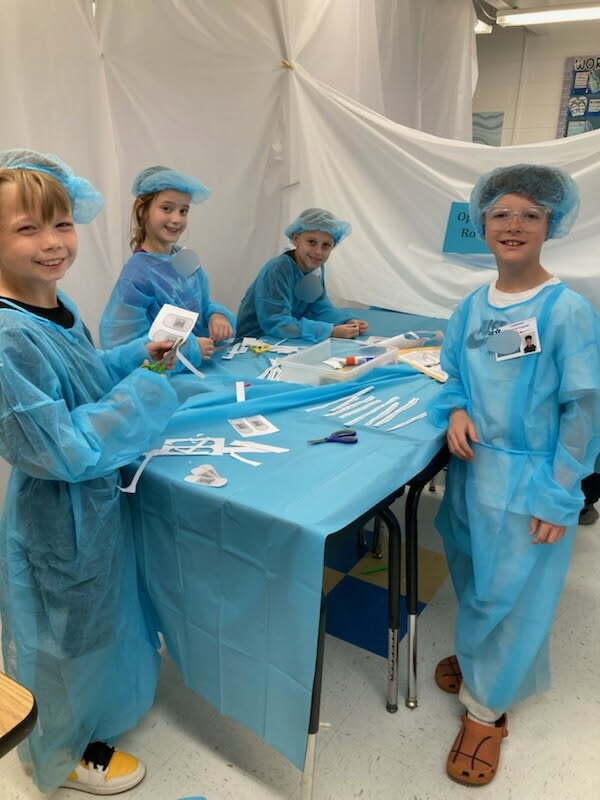 Four students, all wearing surgical gowns, are smiling for a picture while standing at their desks that have been pushed together and covered with a blue plastic tablecloth to resemble an operating table. On the table are cutout pieces of paper, scissors, and a glue stick. 