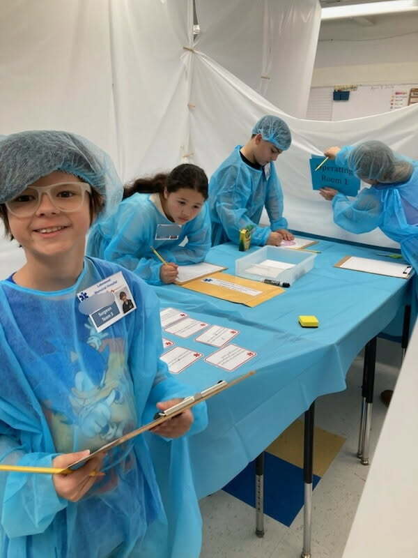 Four students, all wearing surgical gowns, are inside of their makeshift operating room in their classroom. Their desks have been pushed together and covered with a plastic blue tablecloth to resemble an operating table. The student closest to the camera is smiling for a picture. In the background, the other three students are working on their book dissection lesson. 