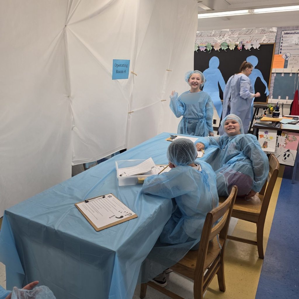 Three students, all wearing surgical gowns, are smiling for a picture while standing at their desks that have been pushed together and covered with a blue plastic tablecloth to resemble an operating table. On the table are cutout pieces of paper, scissors, and a glue stick. 