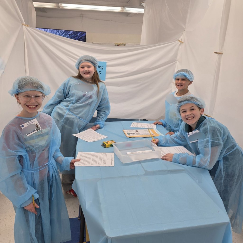 Four students, all wearing surgical gowns, are smiling for a picture while standing at their desks that have been pushed together and covered with a blue plastic tablecloth to resemble an operating table. On the table are cutout pieces of paper, scissors, and a glue stick. 