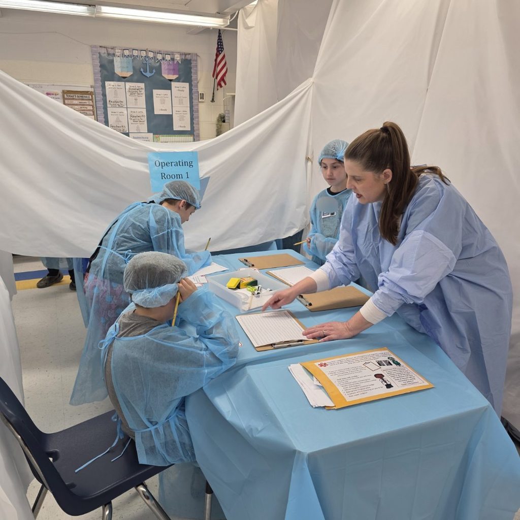 Three students and one teacher are wearing surgical gowns and are working in operating room one. The operating room has been made out of white sheets hanging from the ceiling in the classroom.  