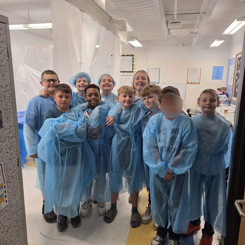 A group of students, all wearing surgical gowns, are standing together for a picture in their classroom. 