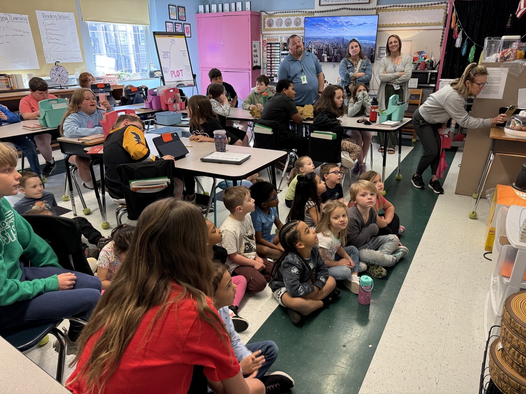 A classroom full of students and teachers watching a TV screen that is not pictured. Some students are sitting on the floor and others are sitting at their desks. 