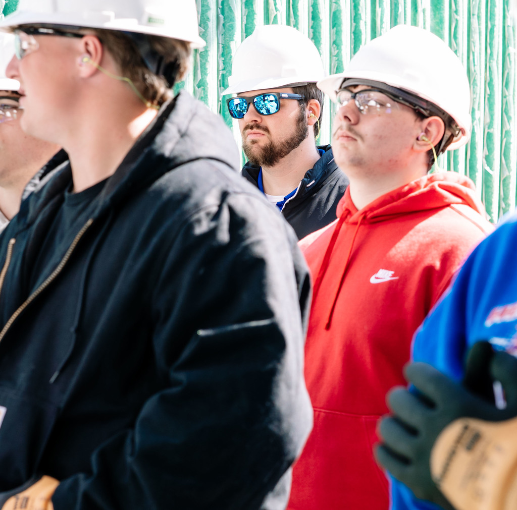 One student, wearing dark sunglasses, listens in during the DOW Tour. He is also wearing a hard hat and ear plugs. Other students are gathered around him.
