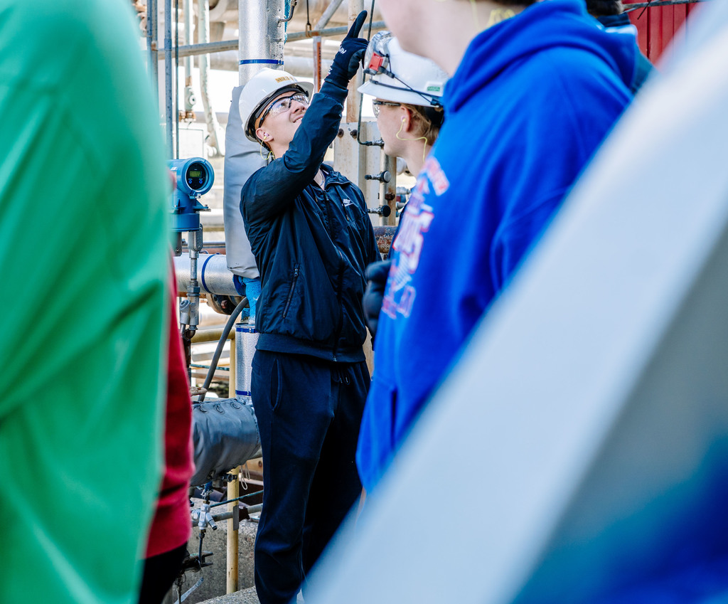 A student on the tour is pointing to a piece of equipment that cannot be seen in the picture. The student is wearing a hard hat, safety glasses, safety gloves, and ear plugs. 