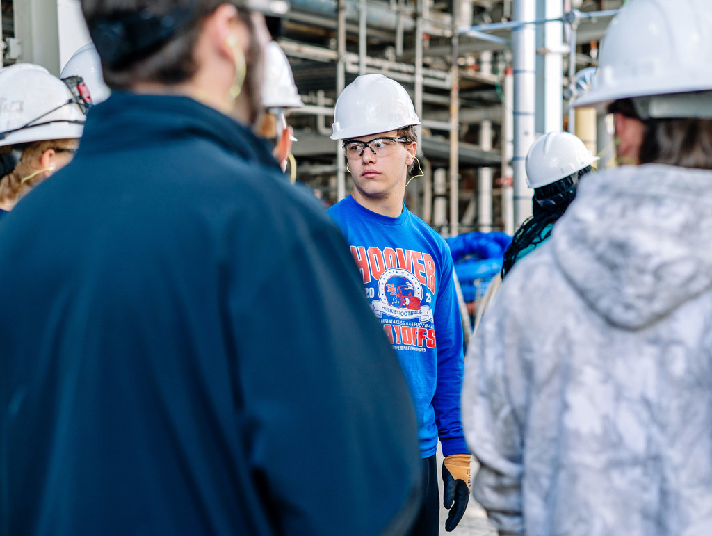 A student is looking at DOW equipment during a tour of the DOW Plant. The student is wearing a hard hat, safety glasses, safety gloves, and ear plugs.