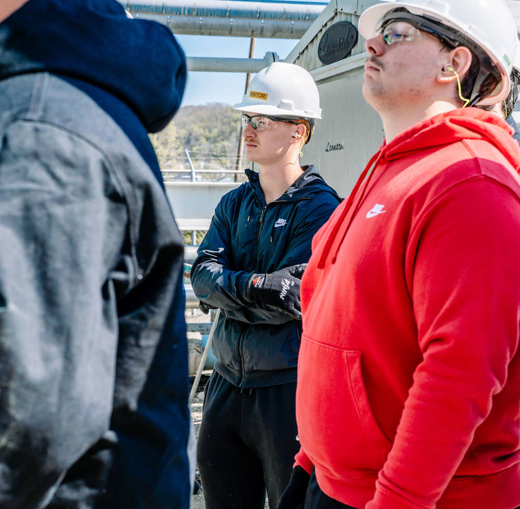 Two students, wearing hard hats, safety glasses, safety gloves, and ear plugs are standing near a row of pipes during the DOW tour.