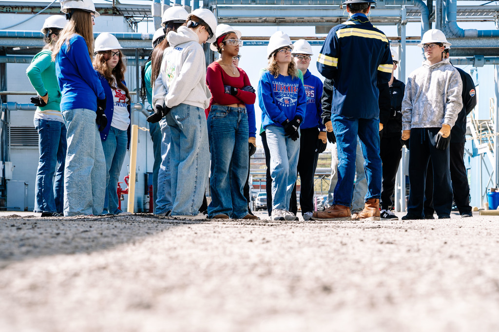 A DOW employee, pictured from behind, is talking to a group of students during the tour of the DOW plant. Students are standing in a semi circle around the employee to listen to the employee. The students and employee are and wearing hard hats, safety gloves, safety glasses, and ear plugs. 