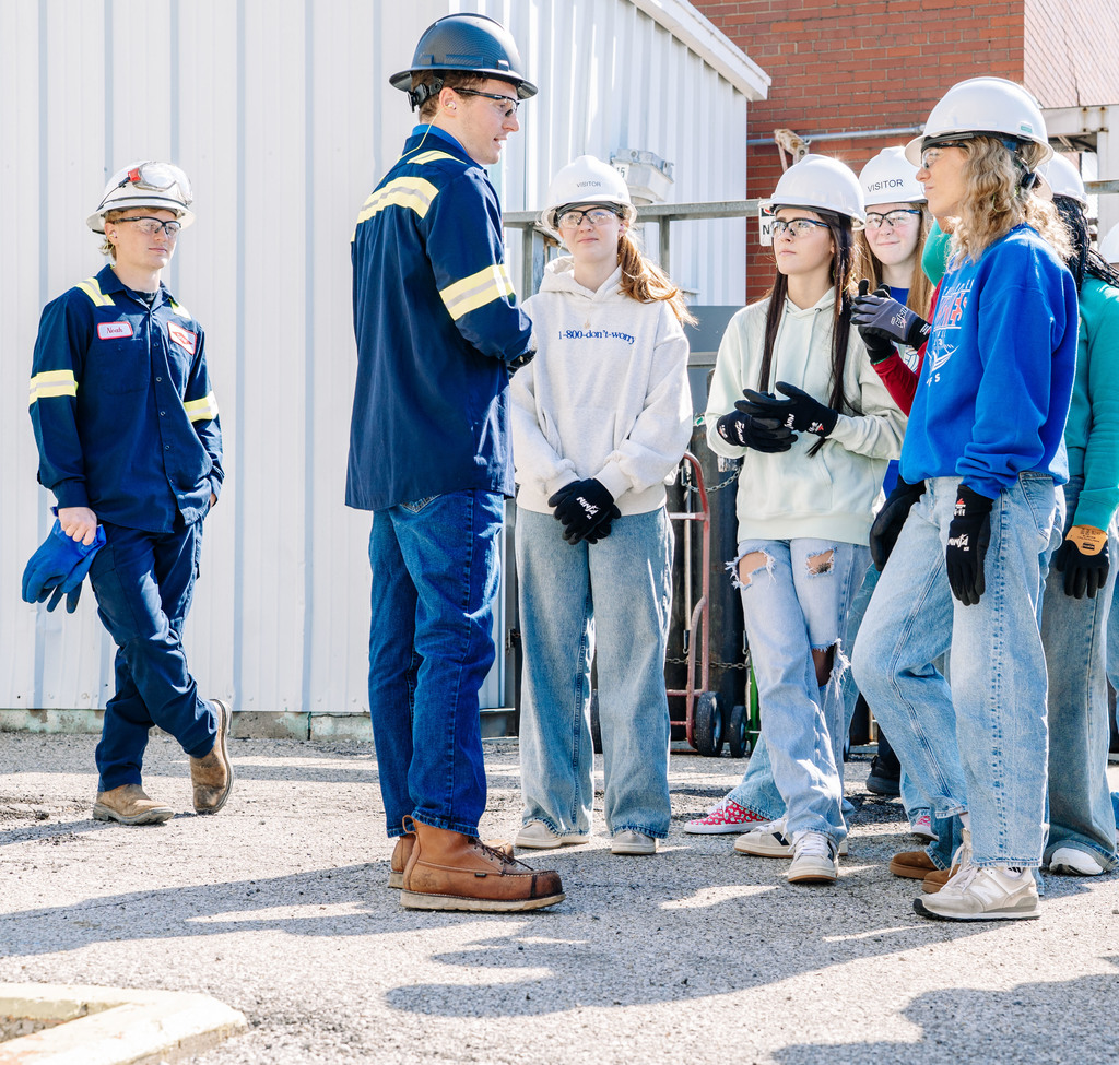 Two DOW employees are standing in front of students during the tour. One employee is directly in front of students, who are standing in a semi circle. The second employee is off to the side of the students and is holding a pair of blue gloves. The students and employees are wearing hard hats, safety glasses, safety gloves, and ear plugs. 
