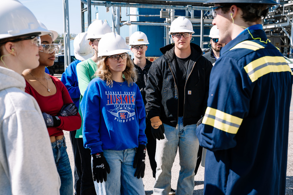 Students are standing in front of a DOW employee during the tour of the plant. Students and the employee are wearing hard hats, safety glasses, safety gloves, and ear plugs. Behind the students are pipelines that run through the plant as well as a blue building. 