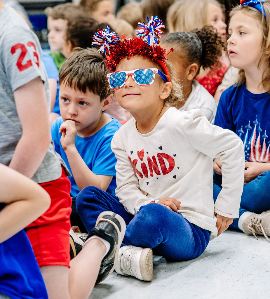A young student is sitting down waiting for the event to start. She is wearing sun glasses that have the American Flag stars on the lenses. She is also wearing a headband that is covered in shiny red little streamers, and on the top have red, white, and blue pom poms and red stars. 