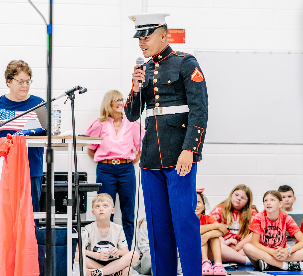 A Marine, in his dress blues, is speaking into a microphone to the Weberwood students during the event.