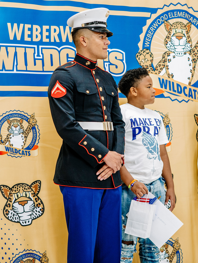 A Marine, in his dress blues, is standing beside his brother who is a student at Weberwood Elementary. The Weberwood student is holding his gift and certificate. They are standing in front of a Weberwood photo banner.
