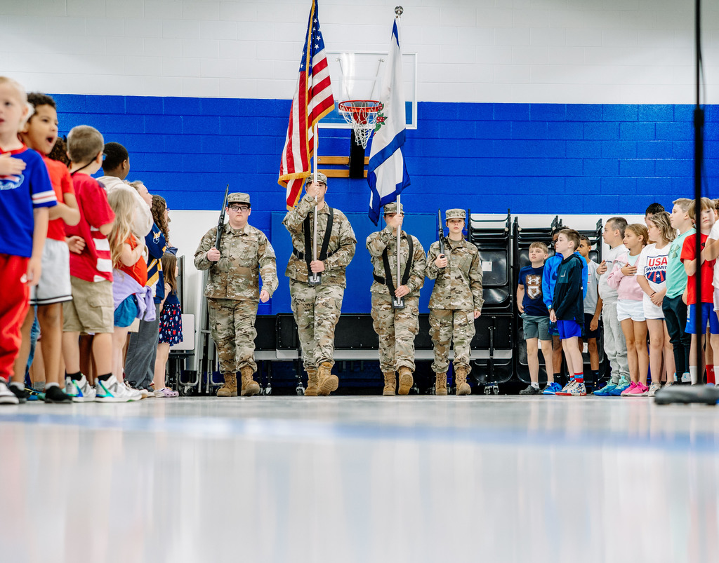 Four members of the Nitro High School ROTC are presenting the colors during the event. The students are on either side of the ROTC students as they march down the cafeteria. In the middle, one ROTC student is holding the American Flag and the other is holding the state flag of West Virginia. On the far left and right, the ROTC students are holding non working air rifles.