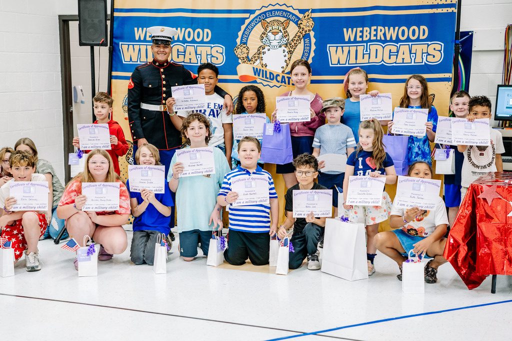 Weberwood students with family members active in the military are standing in front of a Weberwood picture banner. The students are holding a certificate that received during the event. The Marine in his dress blues is standing beside his brother, who is a Weberwood student.