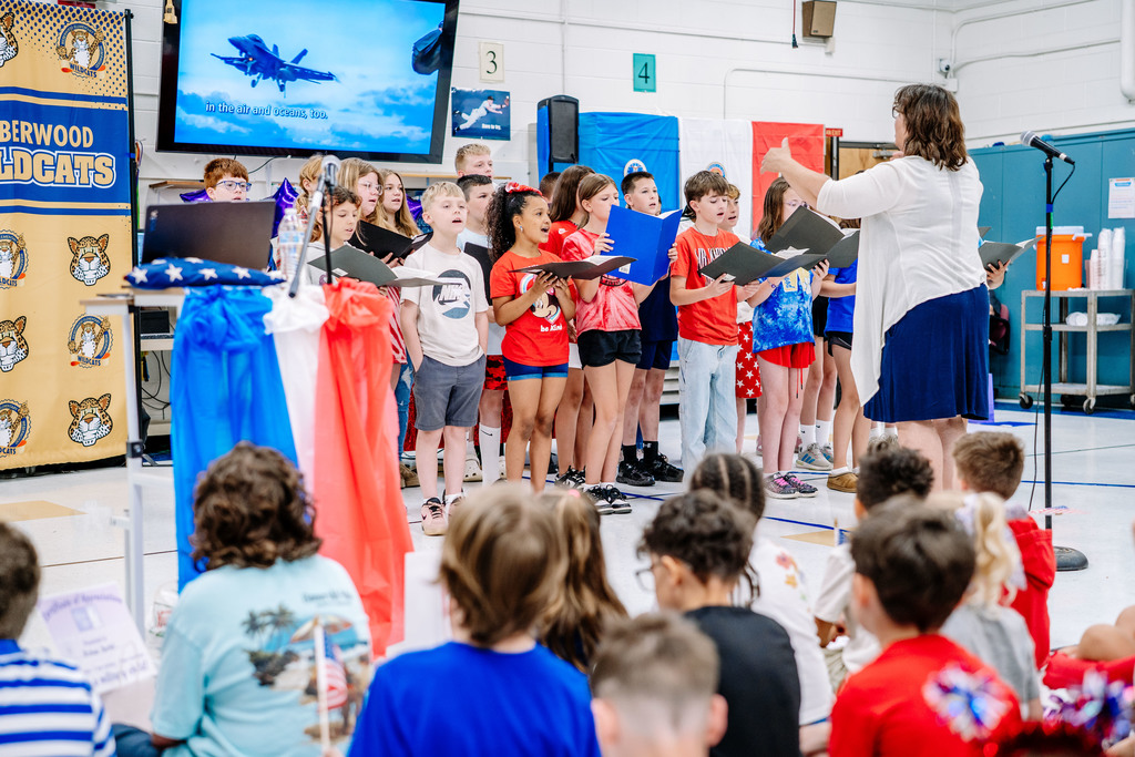 The Weberwood Elementary choir is standing in the front of the cafeteria signing a song during the event. The choir director is leading the choir. Behind the choir is a TV with the words of the song and a military jet.