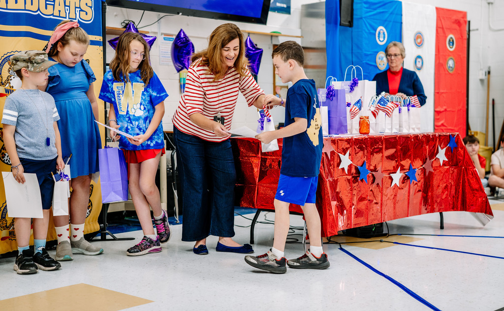 A Weberwood student with a family member in the military is receiving his gift from a teacher. The teacher is smiling and the student is holding a small white bag in one hand and his certificate in the other hand.