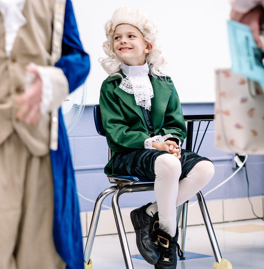 A student, dressed as Thomas Jefferson, sitting in his chair during the wax museum. He is looking off to his right and smiling. 