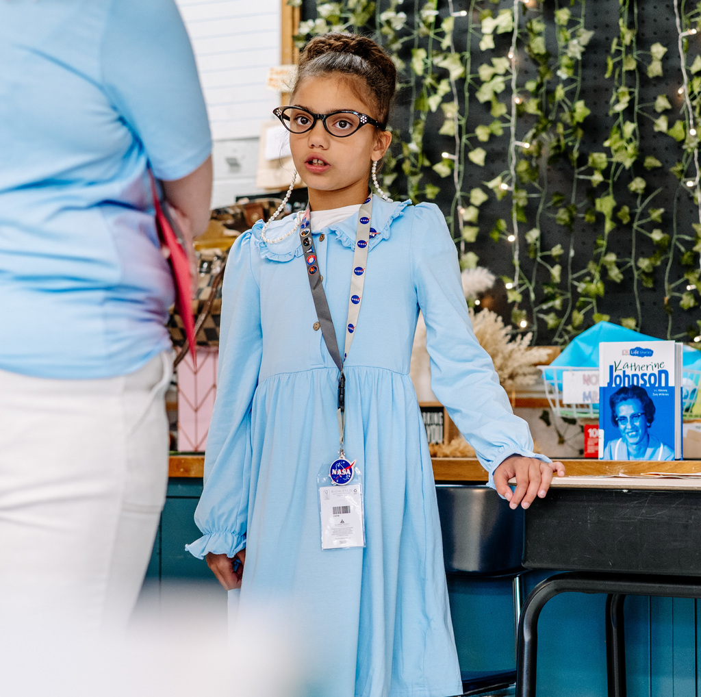 A student dressed as Katherine Johnson is standing with her hand on her desk giving her presentation. Behind her is a book about Katherine Johnson. 
