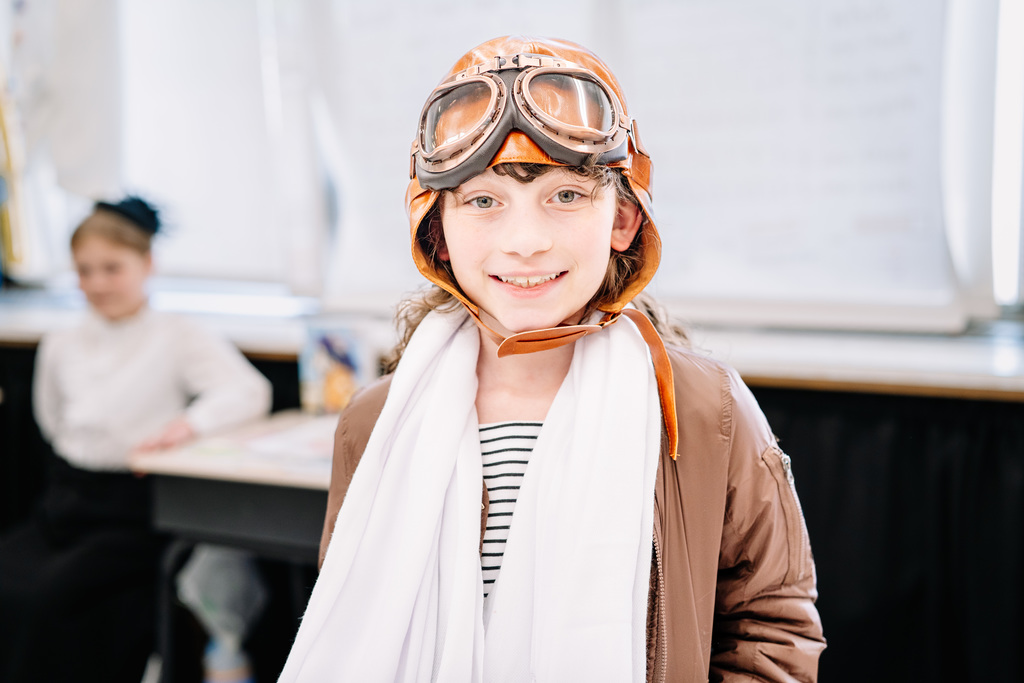 A student dressed as Amelia Earhart is smiling for a picture. She has on a leather aviation cap, aviation goggles, and his wearing a long white scarf.