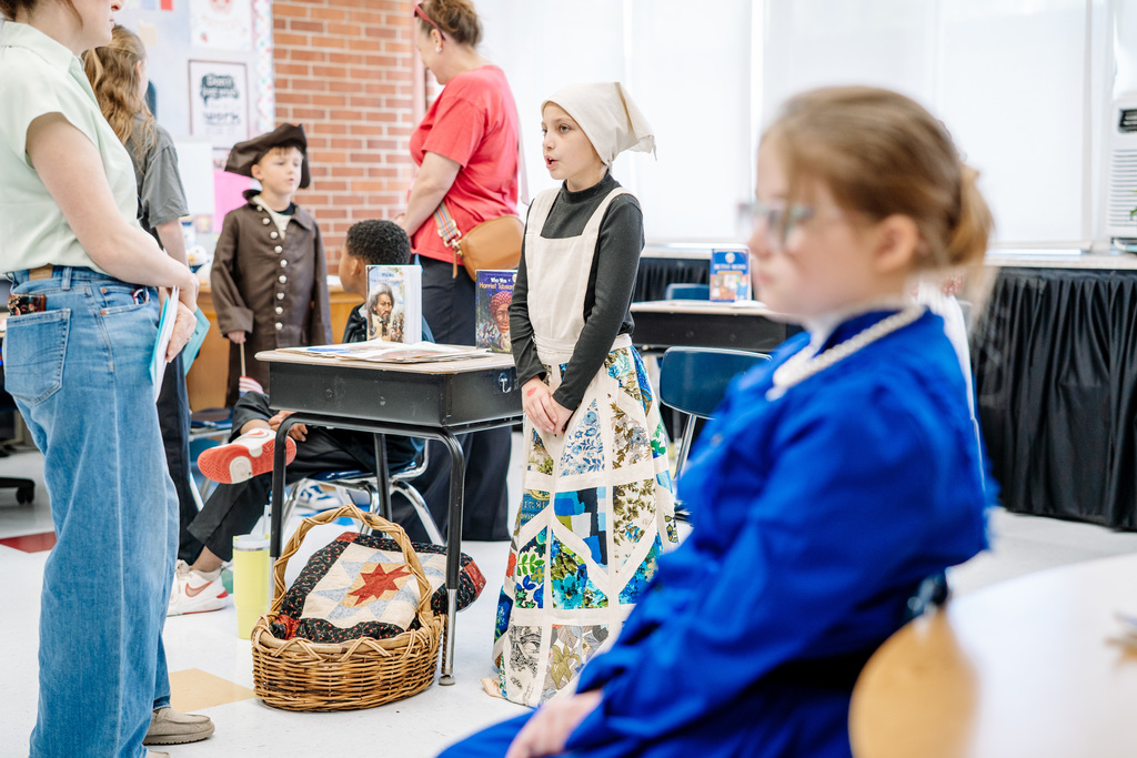 A student standing at her desk giving her presentation about Harriet Tubman. She is wearing a had covering and floor length quilted dress. 