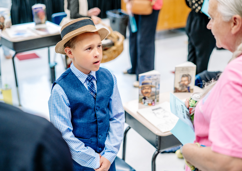 A student dressed as Milton Hershey standing up at his desk speaking to a woman listening to his presentation.