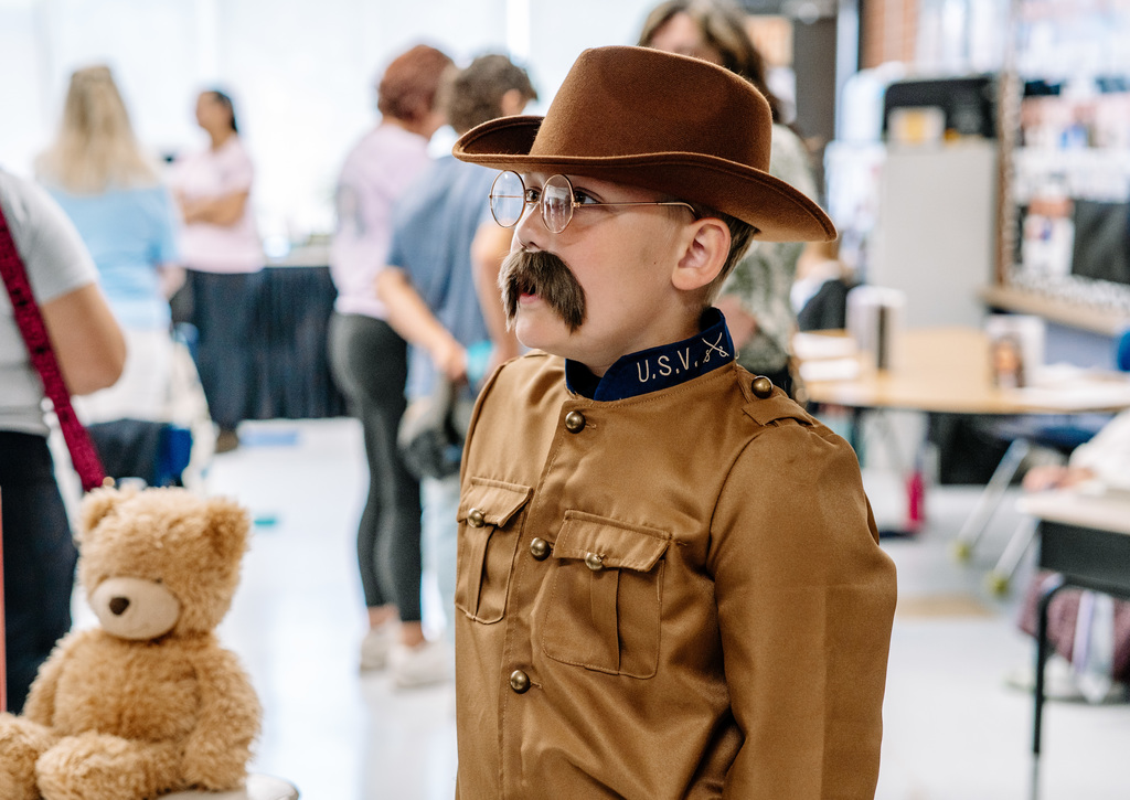 A student dressed as Teddy Roosevelt in his military uniform. The student also has on a fake mustache to mimc Roosevelt's mustache. Beside the student is a teddy bear.