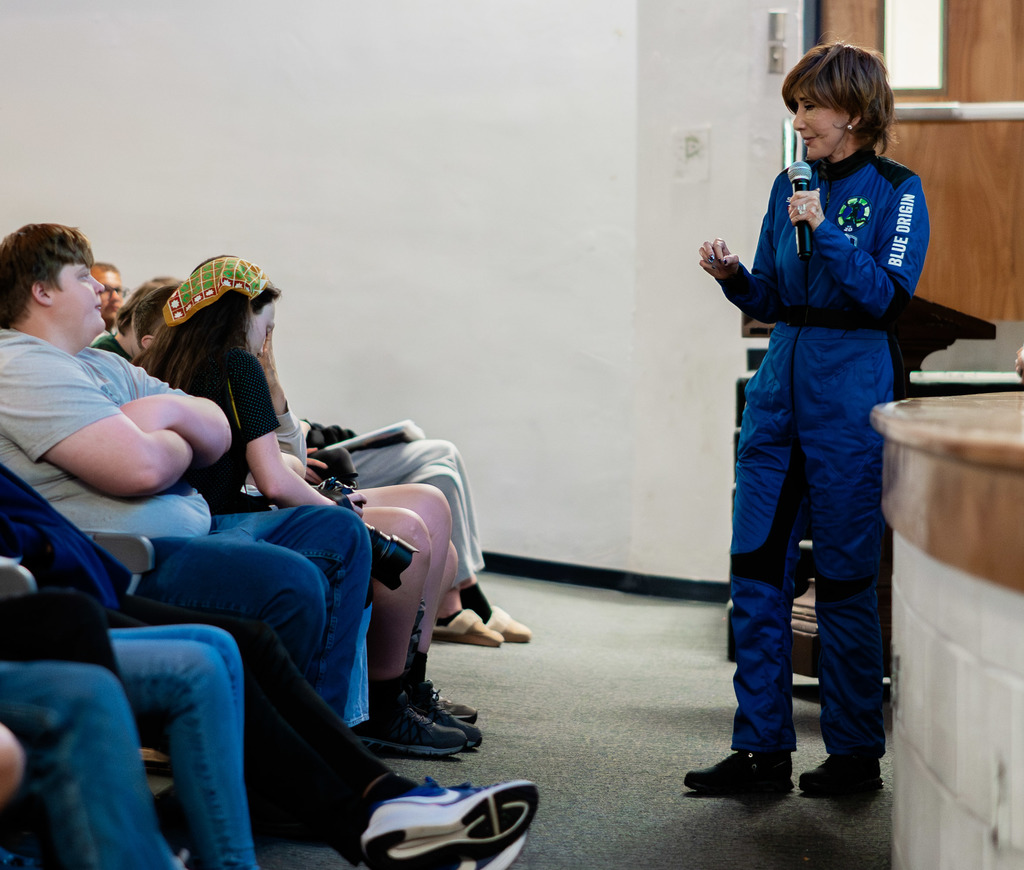 Sharon Hagle, wearing her Blue Origin space suit, is standing at the front of the SAHS auditorium talking to the student body. 