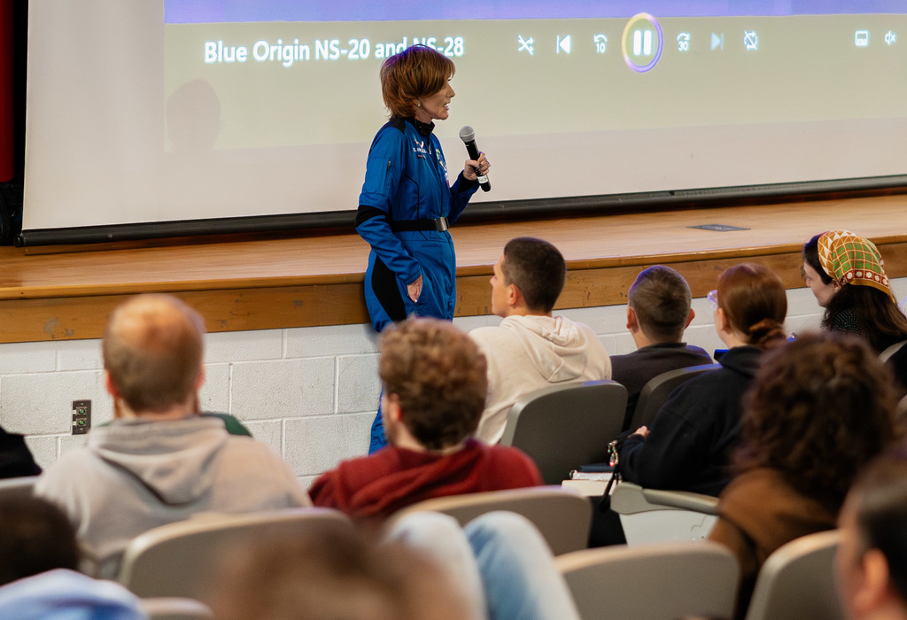 Sharon Hagle, wearing her Blue Origin space suit, is standing at the front of the SAHS auditorium talking to the student body. Behind her is a projector that is playing her presentation. 