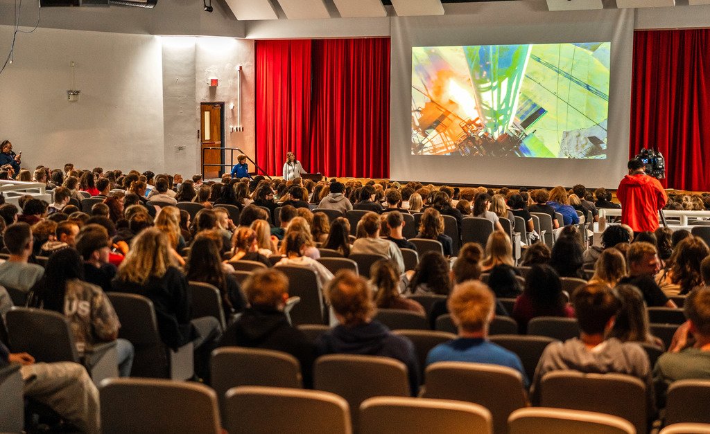 Students in the auditorium watching a video on a large projection screen of Sharon's space shuttle take off. You can see fire coming out of the bottom of the  space craft as it launches. 