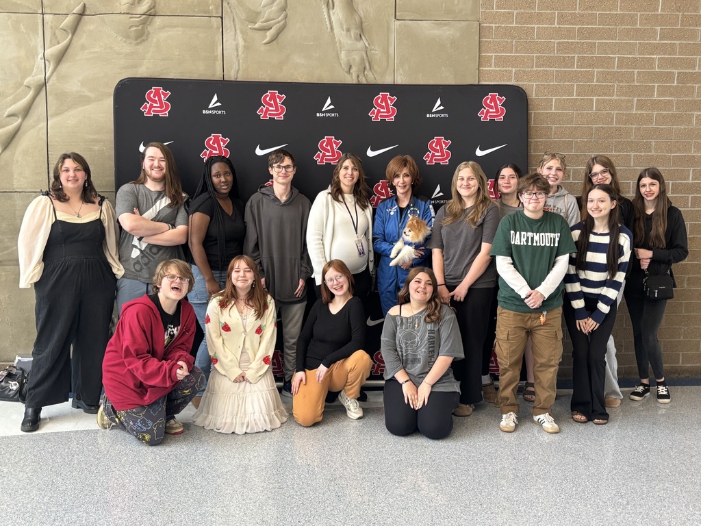A group photo of SAHS students, the SAHS Principal, and SAHS Alumni Sharon Hagle. The group is posing in front of the SA photo backdrop which is black, with white Nike checkmarks and the Red SAHS logo.