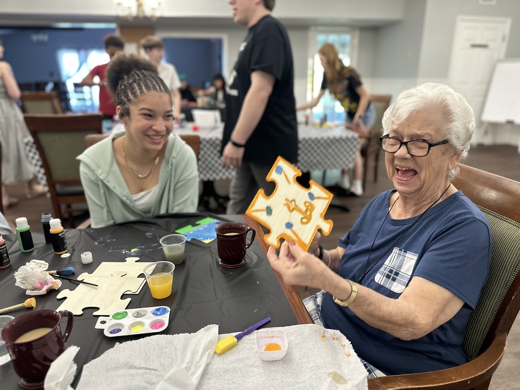 A nursing home resident, sitting at a table, is holding up her finished painting project and laughing. A student is sitting beside her and looking at her and smiling. 