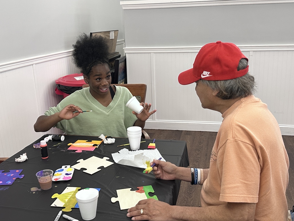 A student and a nursing home resident are sitting at a table doing a painting project. The student is in the middle of talking to the nursing home resident. 